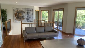 A modern living room with a grey sofa, wooden flooring, a large abstract painting, and a view of the outdoors through large windows. - Grand Retreat in Mount Eliza (Mount Eliza)