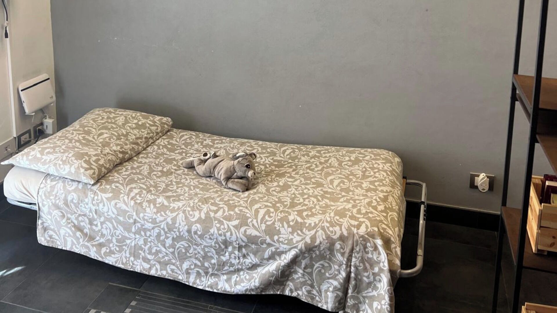 Un lit simple avec une couvre-lit à motifs et des oreillers, un petit éléphant en peluche et une table de chevet avec une lampe.