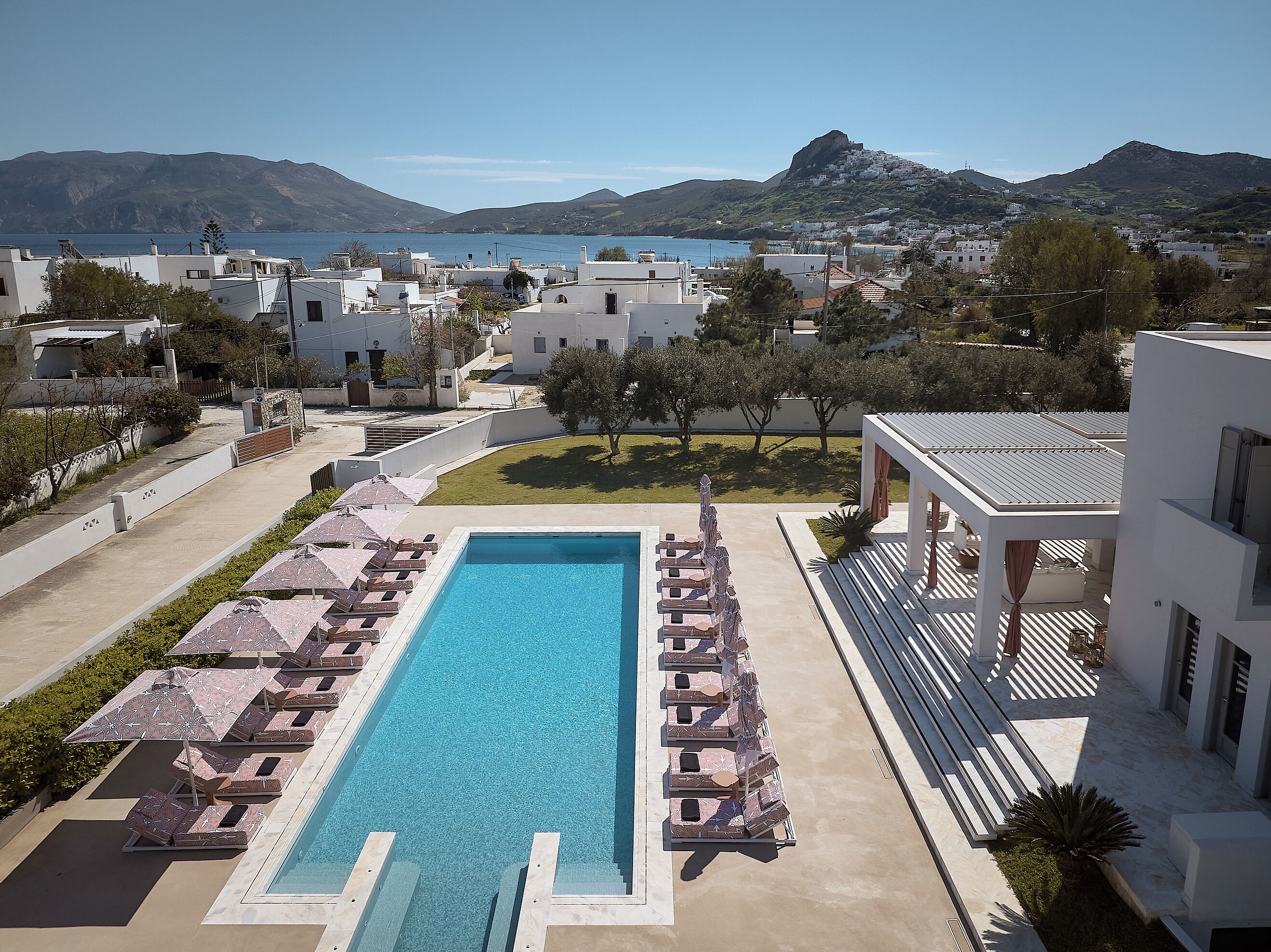 En swimmingpool med solstolar och parasoller, omgiven av byggnader och med utsikt över havet och bergen.