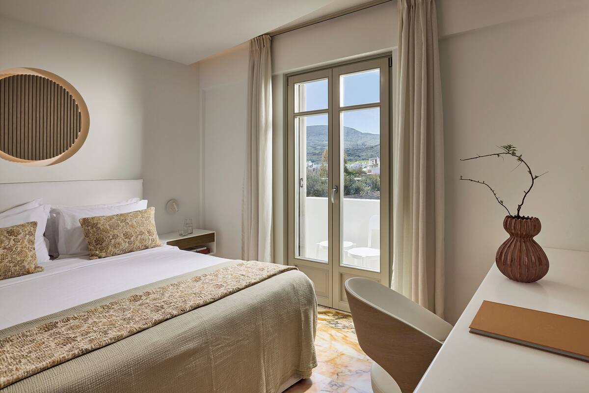 Ett hotellrum med en säng, ett skrivbord, en stol, ett fönster med utsikt över omgivningarna och en dekorativ vas.