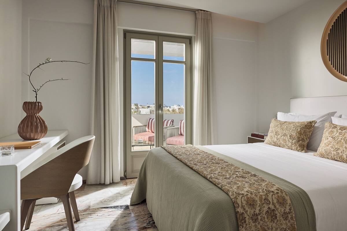 Ett hotellrum med en säng, ett skrivbord, en stol, en vas och utsikt över omgivningarna.
