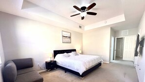 Une chambre à coucher comprenant un lit, une table de chevet, une chaise, un ventilateur de plafond et une porte donnant sur une autre pièce.