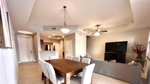 Un grand espace à manger avec une table en bois et des chaises blanches, un ventilateur de plafond et une cuisine en arrière-plan.