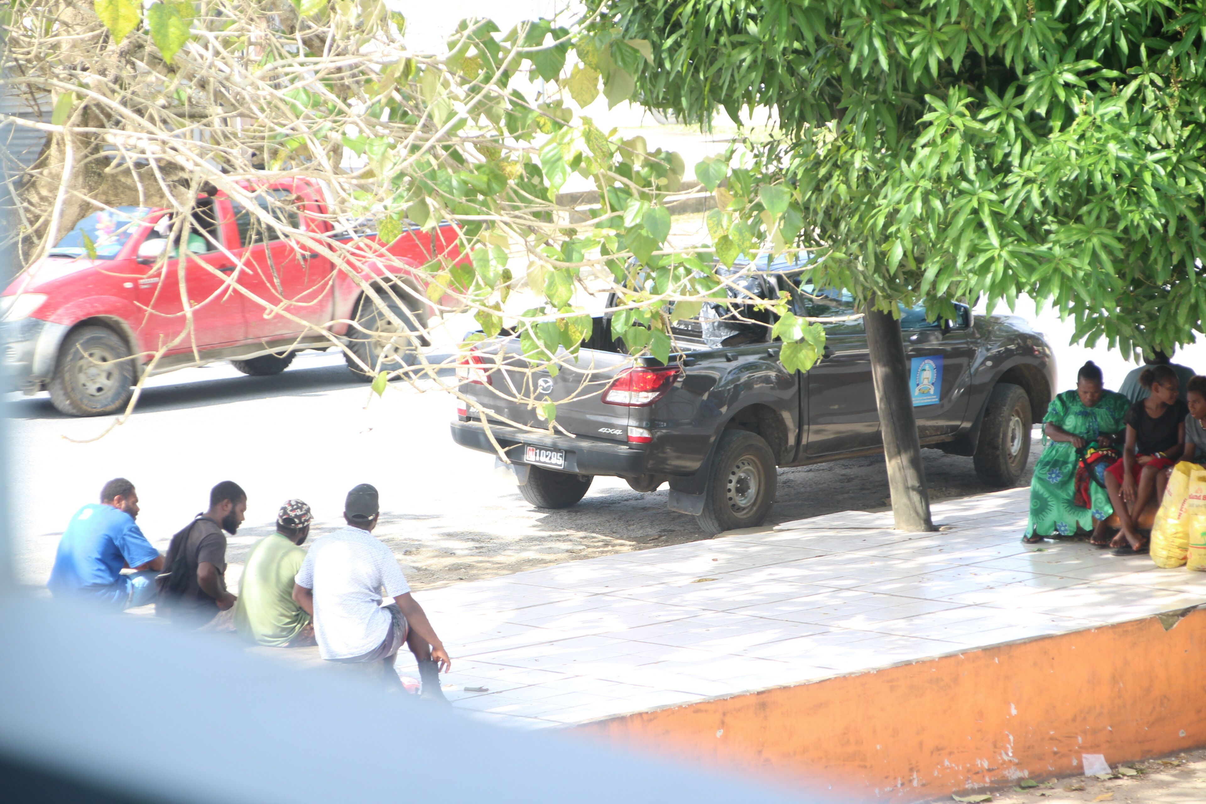 Un grupo de personas sentadas en una acera junto a un árbol y coches aparcados.