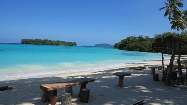 A beach with a wooden bench and a small table, surrounded by palm trees and clear turquoise water. - Sunshine Inn (Luganville)