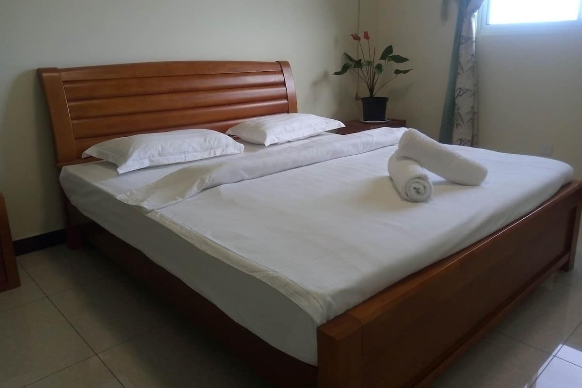 A neatly made bed with white linens and a rolled towel, a wooden headboard, and a potted plant on a bedside table.