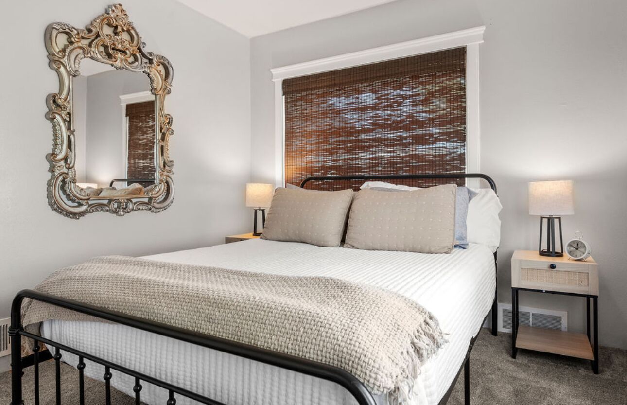 A bedroom with a bed, two bedside tables, a mirror, and a window with blinds.