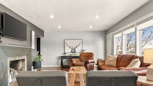 A modern living room with a fireplace, leather sofa, and a large window offering a view of the outdoors. - Riverview Home - walking trails - convenient location (Clarkston)