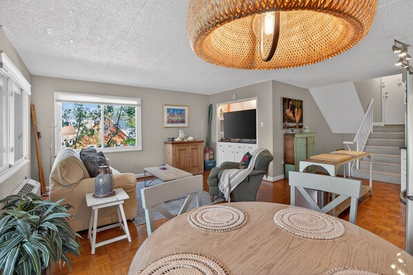 A spacious living area with a large window, a dining table with woven placemats, a sofa, a chair, and a staircase leading to an upper level.