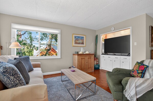 A living room with a sofa, a coffee table, a TV, and a window with a view of trees. - Downtown Rehoboth Beach Condo Sleeps 4 Parking (Rehoboth Beach)