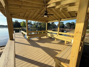 Un gazebo in legno con ventilatore a soffitto, panche di legno e vista su un lago e case sullo sfondo.