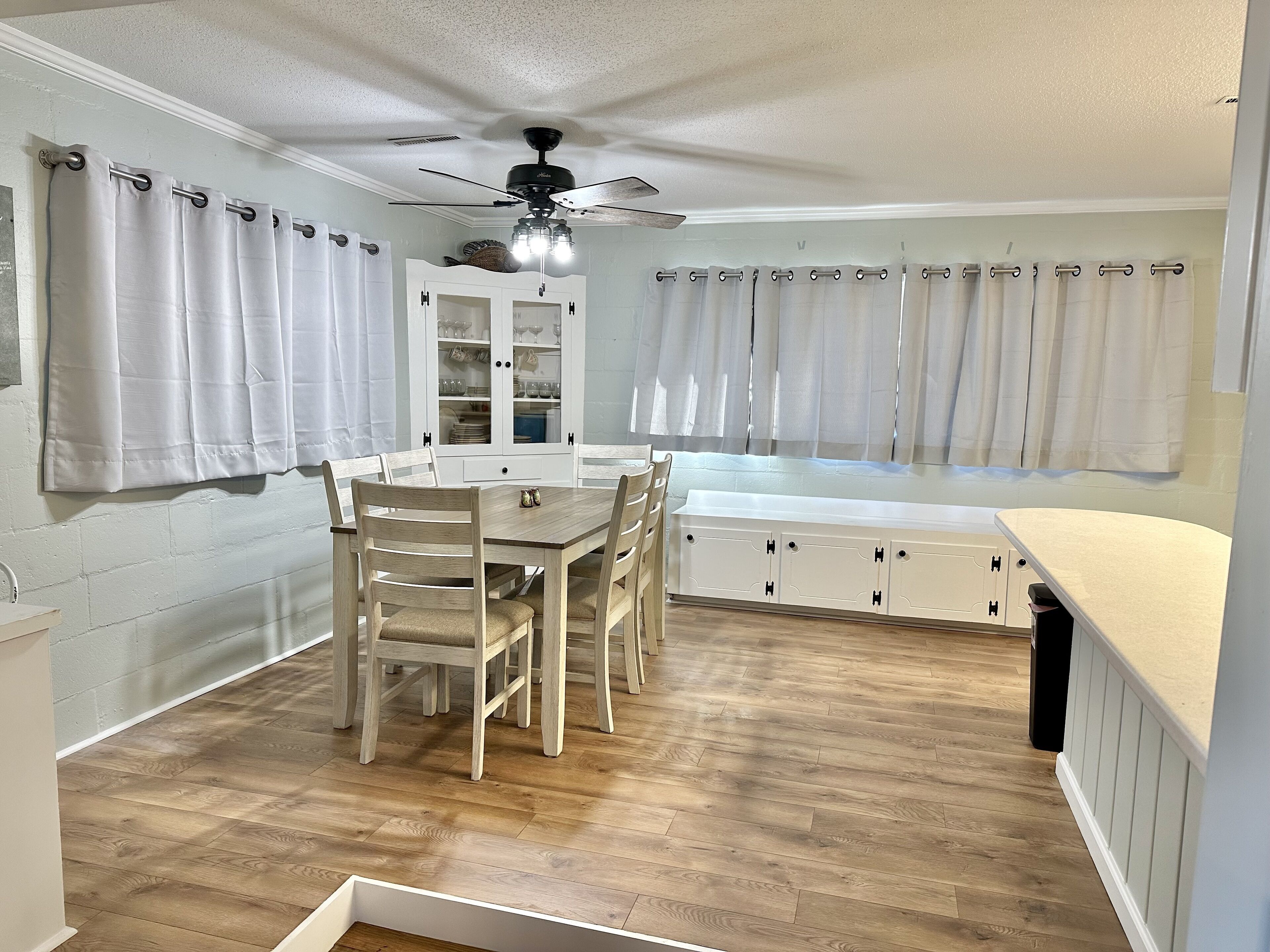 A kitchen with wooden flooring, white cabinets, a dining table with chairs, and a ceiling fan.