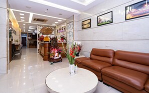 A modern lobby with a brown leather sofa, a round white table with a vase of flowers, and a reception desk with a sign. - Hotel Sao Mai (Ho Chi Minh City)