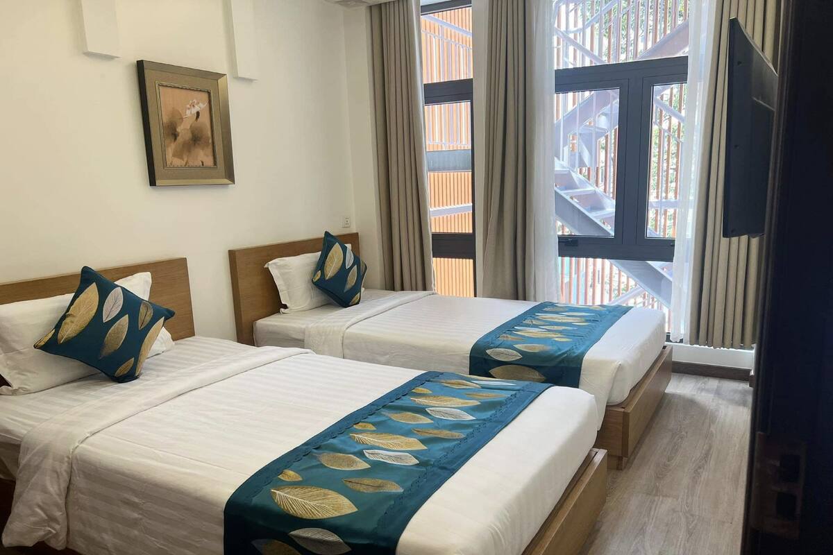 A hotel room with two beds, each with a blue and white bedspread, a framed picture on the wall, and a window with a view of a wooden structure outside.