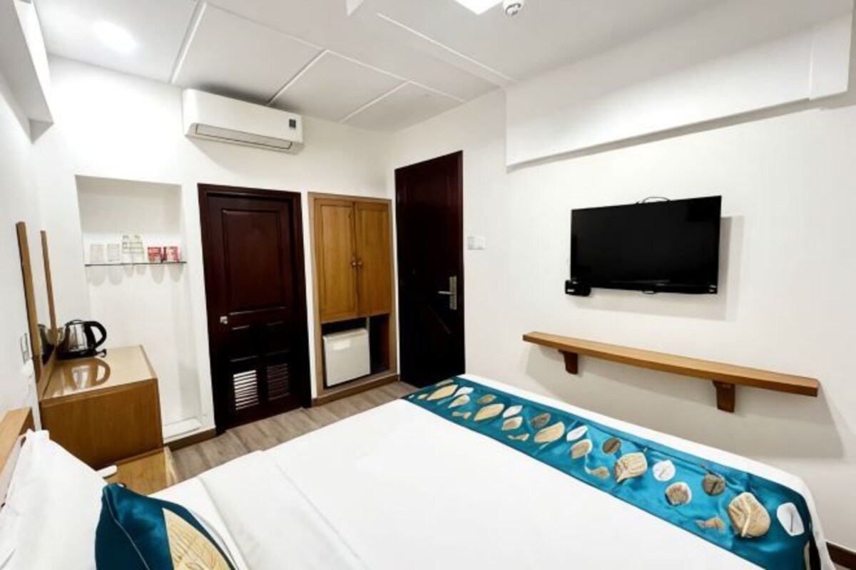 Habitación de hotel con cama, televisor, estante, armario y escritorio con hervidor.