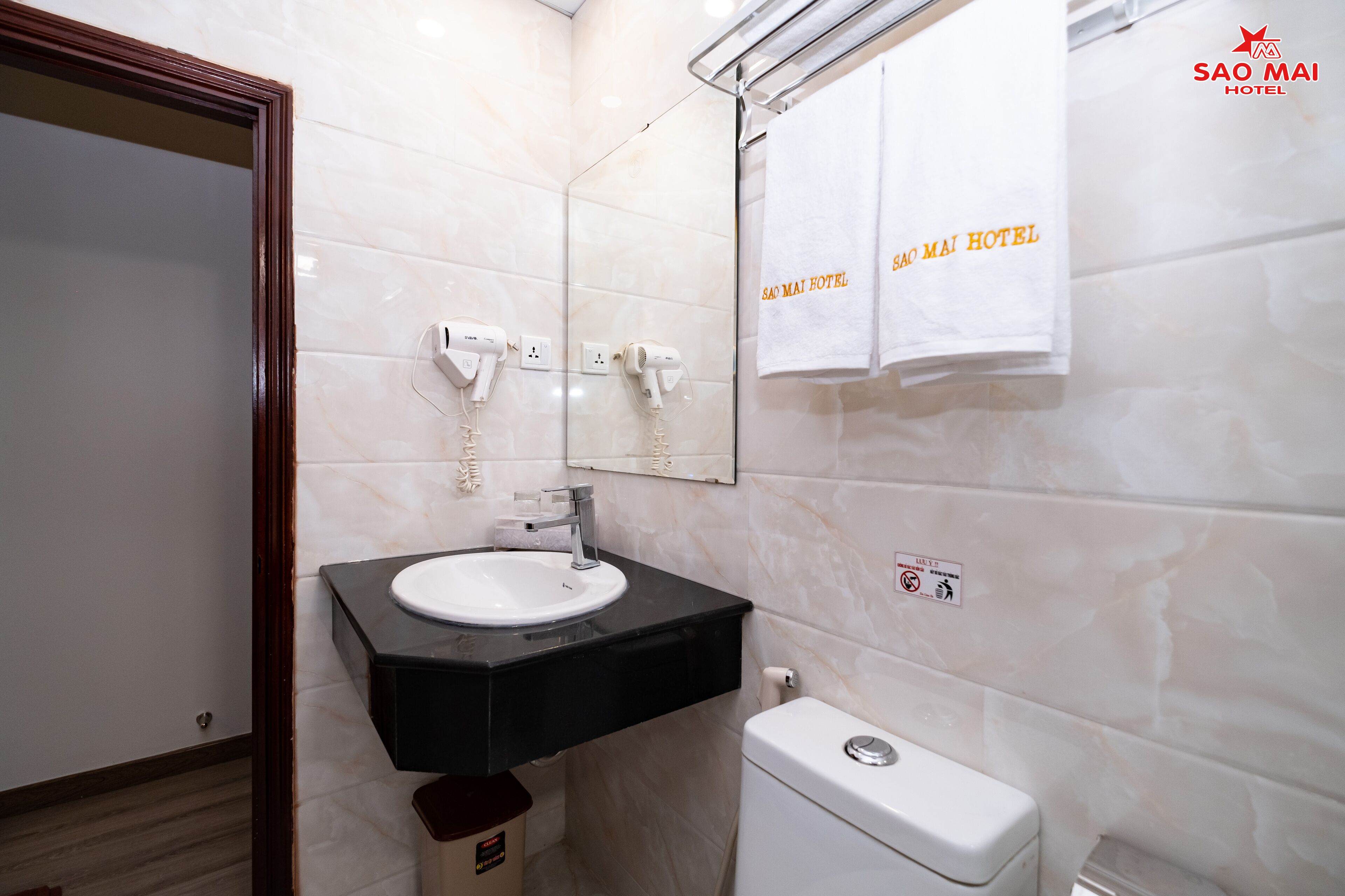 Un baño moderno con un lavamanos blanco, encimera negra y un inodoro. Hay dos secadores de pelo montados en la pared, y un perchero con toallas colgando. Las paredes están revestidas con baldosas de color claro, y hay un espejo sobre el lavamanos.