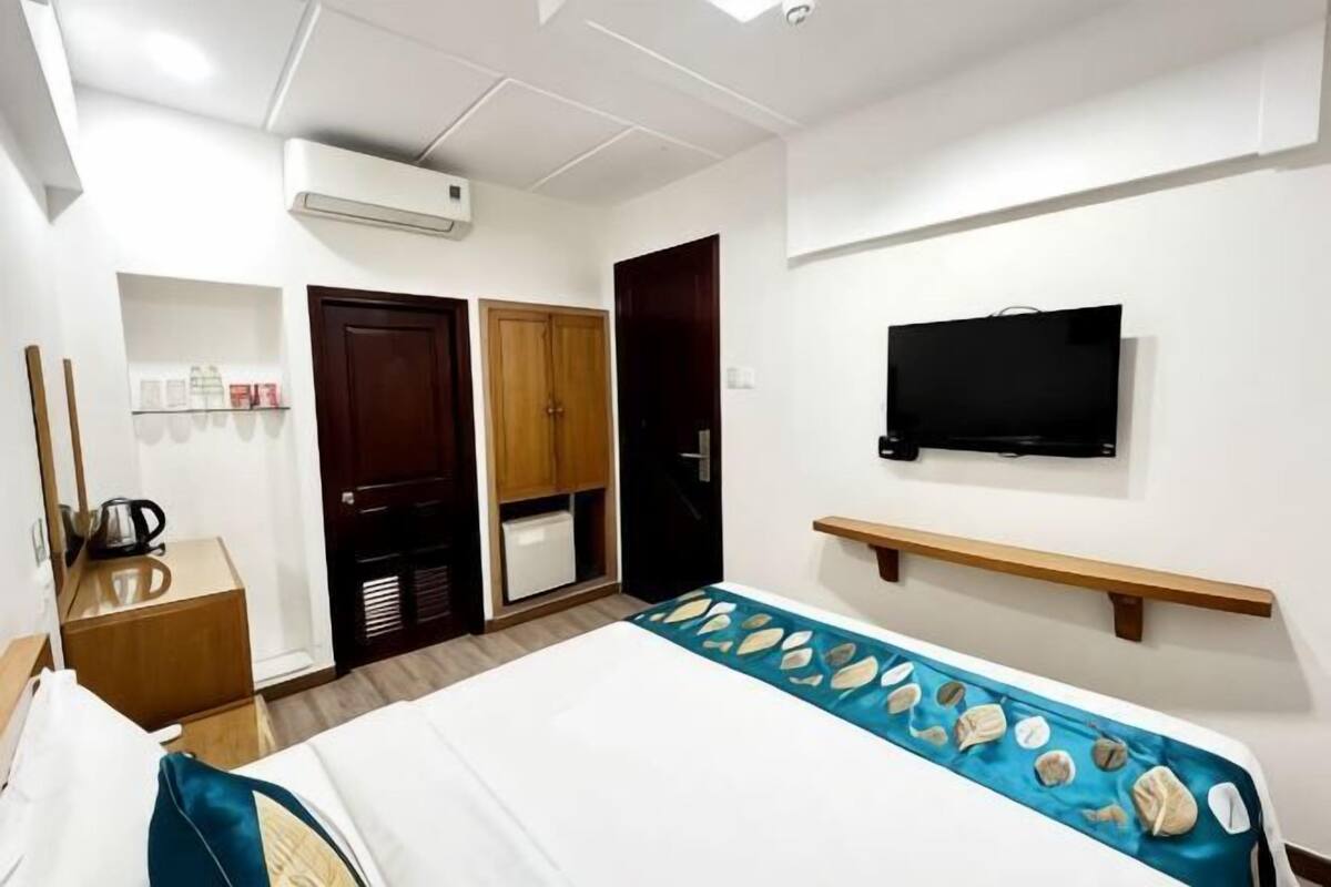 A hotel room with a bed, a TV, a shelf, a wardrobe, and a desk with a kettle.
