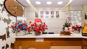Zona de recepción de hotel con un mostrador de madera, un cartel que dice 'Recepción' y varios arreglos florales.