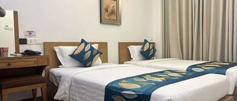A hotel room with two beds, a desk, and a chair. There is a painting on the wall and a window with curtains.