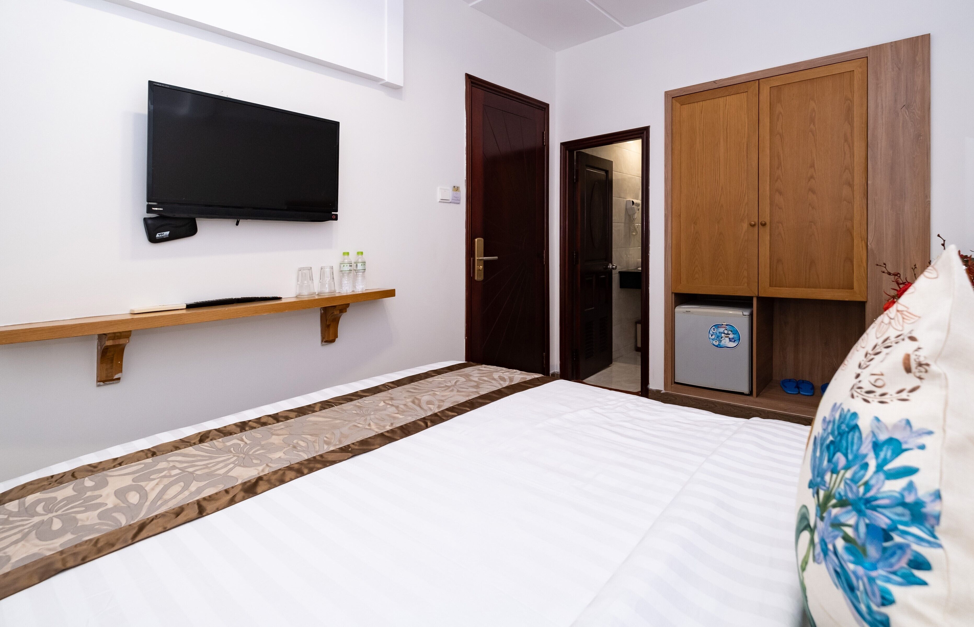 A hotel room with a bed, a TV mounted on the wall, a wooden wardrobe, and a small shelf with toiletries.