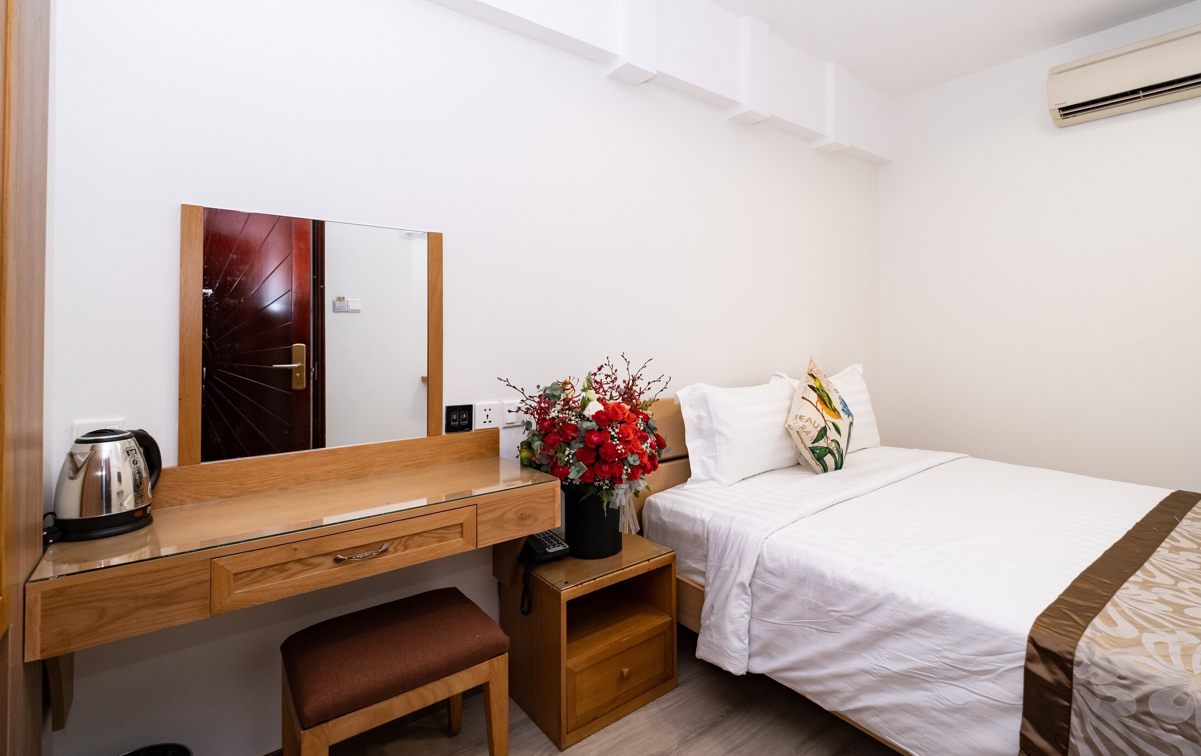 Camera d'albergo con una scrivania in legno, uno specchio, un letto con lenzuola bianche, un comodino con un vaso di fiori e uno sgabello.