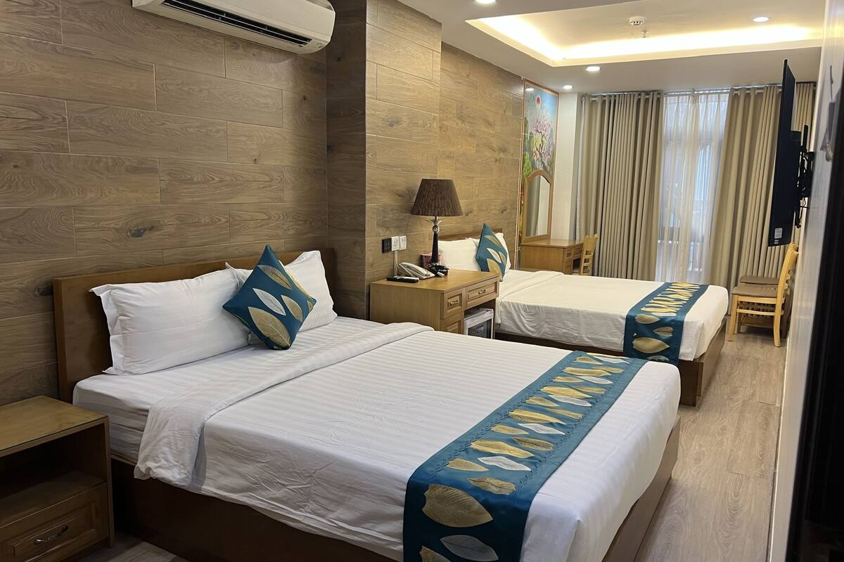 A hotel room with two beds, wooden flooring, a ceiling light, a nightstand, a lamp, a mirror, and a window with curtains.