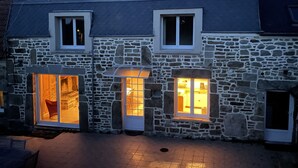 A stone building with two windows and a door, illuminated by warm interior lighting. - L'Odyle - House for 6 people 1 Km from the sea (Fermanville)