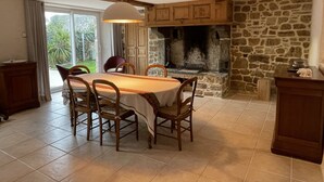 A dining area with a stone fireplace, wooden chairs, a table with a white tablecloth, and a hanging light fixture. - L'Odyle - House for 6 people 1 Km from the sea (Fermanville)