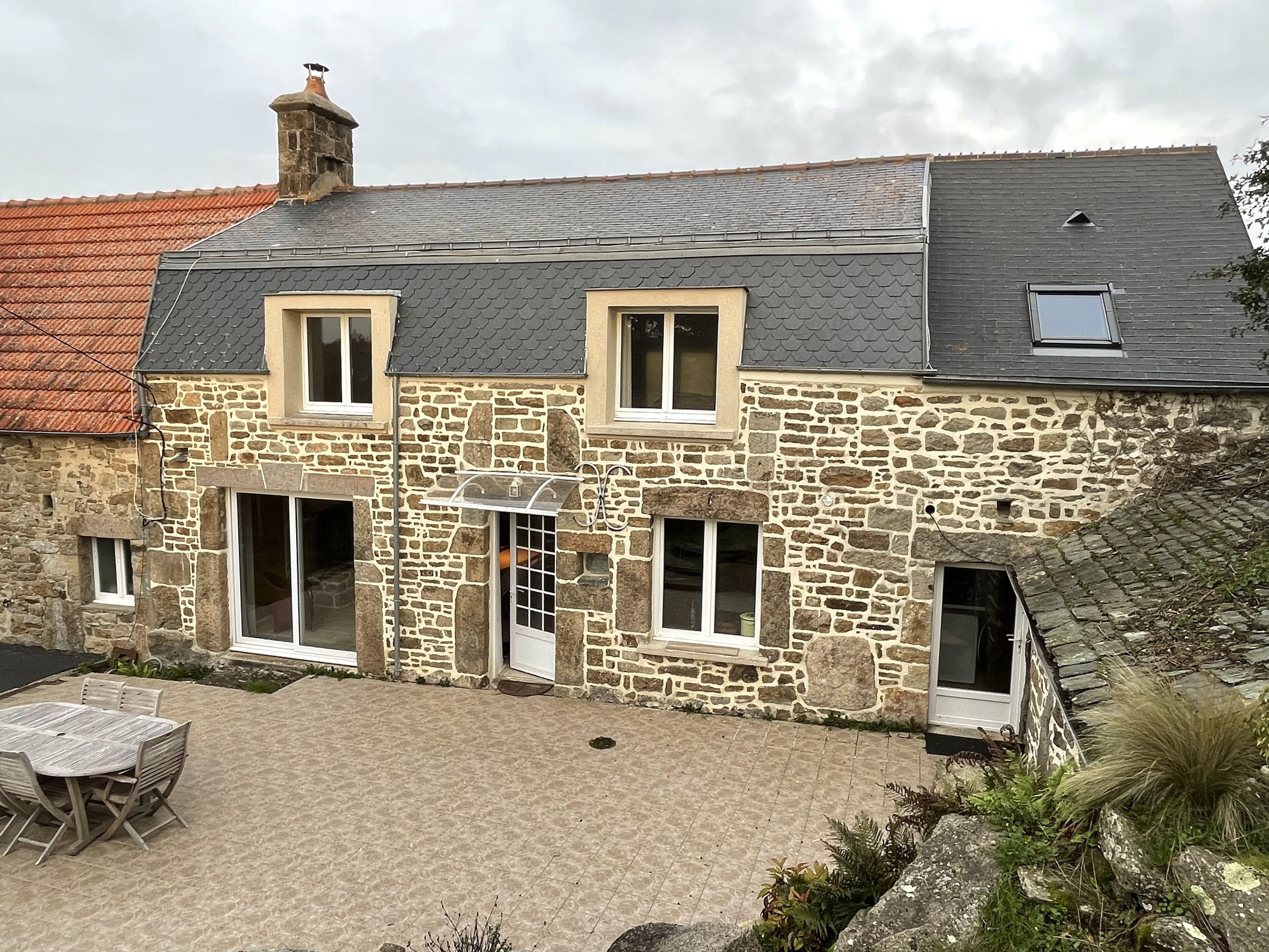 A stone house with a tiled roof, a small balcony, and a paved patio area with a table and chairs.