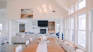 A spacious dining area with a long wooden table and white chairs, a chandelier, and a television above the fireplace. - LUXURY, OCEANFRONT, Pool, ELEV, Rec Rm, Beach Access, Ocean Views! WL983, Fairlight (Corolla)