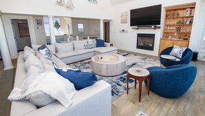 A spacious living room with a large sectional sofa, a blue armchair, a round coffee table, and a fireplace. - LUXURY, OCEANFRONT, Pool, ELEV, Rec Rm, Beach Access, Ocean Views! WL983, Fairlight (Corolla)