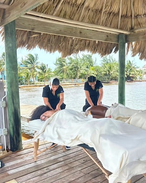A wooden deck with a thatched roof, a massage table, and a massage therapist attending to a client.