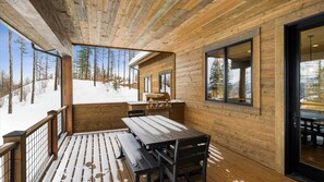 A wooden deck with a grill and picnic table, overlooking a snowy landscape with trees. - The Sunrise Ridge Chalet (Whitefish)
