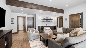 A modern living room with a grey sofa, armchairs, and a coffee table. There is a kitchen area with a sink and cabinets, and a wooden ceiling beam.