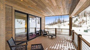 A wooden deck with a glass door leading to a room, outdoor seating, and a snowy mountain view. - The Sunrise Ridge Chalet (Whitefish)