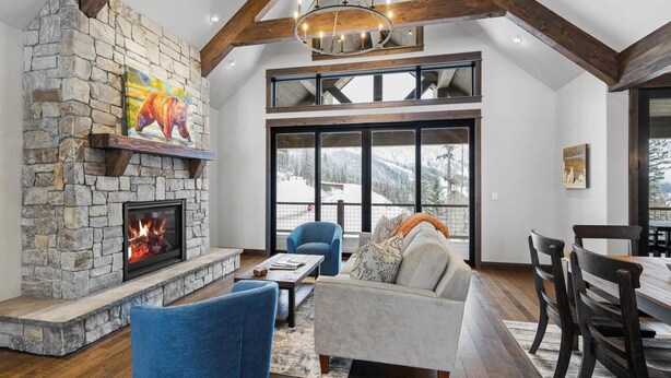 A cozy living room with a stone fireplace, comfortable seating, and large windows offering a view of the snowy outdoors. - The Sunrise Ridge Chalet (Whitefish)