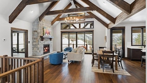 A spacious living area with a stone fireplace, wooden beams, and a large window overlooking a snowy landscape.