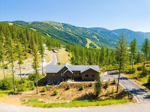 A cabin with a sloped roof, surrounded by a forested area with a winding road leading to it. - The Sunrise Ridge Chalet (Whitefish)