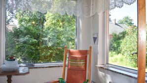 A room with a window overlooking a garden, a wooden chair with a striped cushion, a small table with a candle, and a wooden crate labeled "KUB".