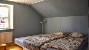 A small, single-room apartment with a sloped ceiling, a window with a view of a building outside, a radiator, a floor lamp, and a bed with patterned bedding.