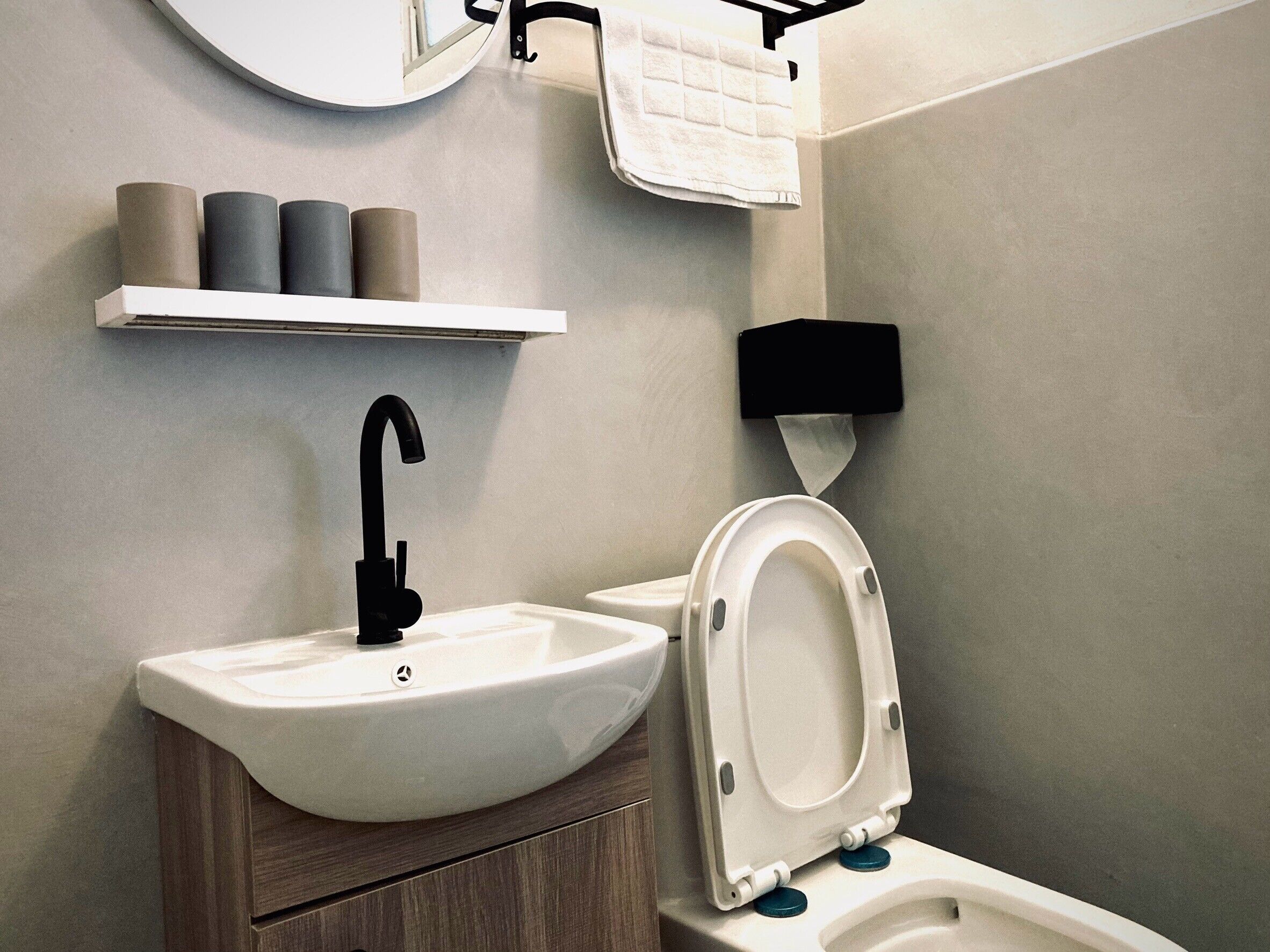 A modern bathroom with a white sink, black faucet, and a toilet with a white seat. There are toilet paper rolls on a shelf above the sink.