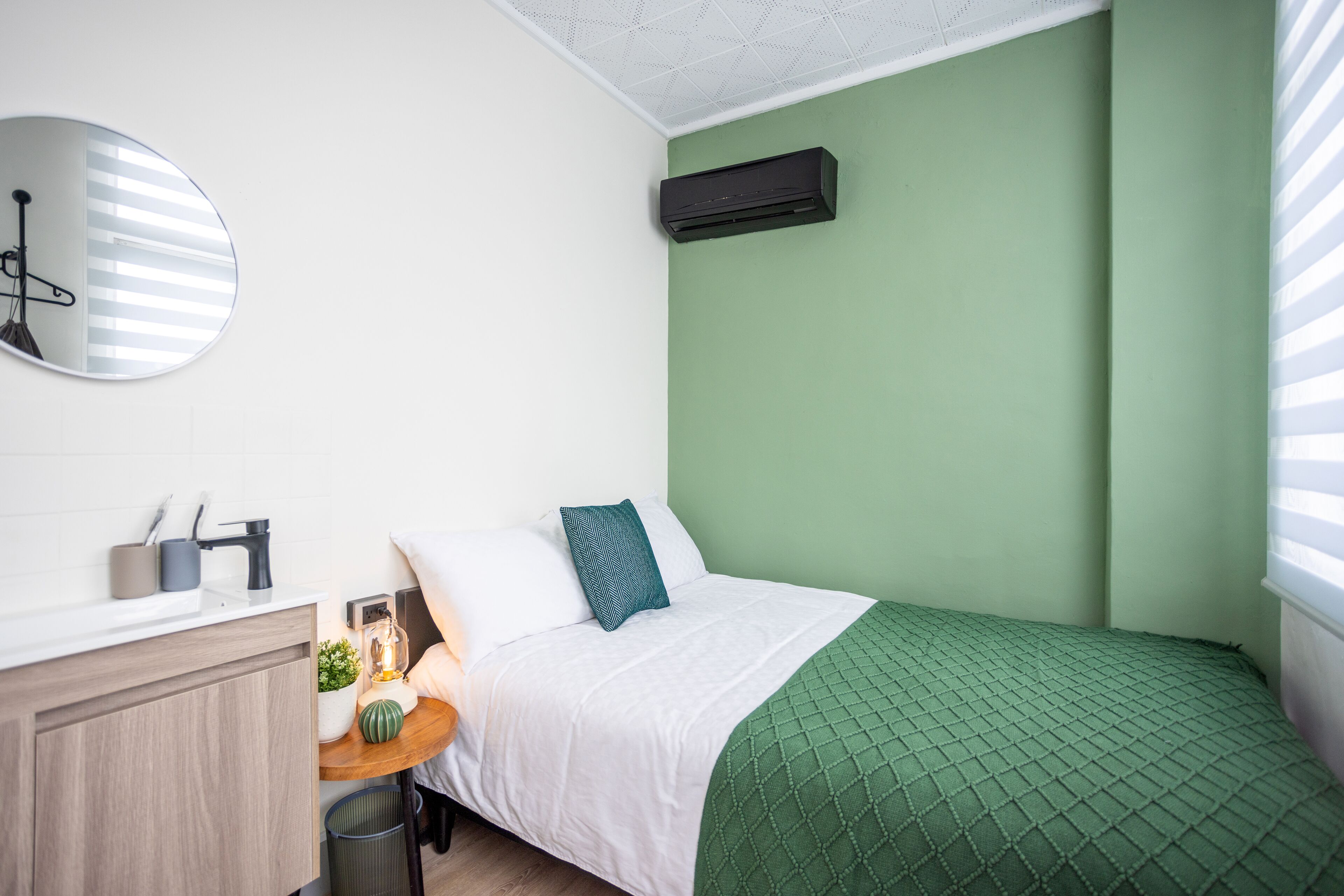 A bedroom with a green wall, a white bed, a round mirror, a bedside table with a lamp, and a small plant.