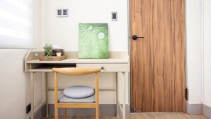A room with a wooden door, a desk with a plant, a cushion, and a wall-mounted light switch.