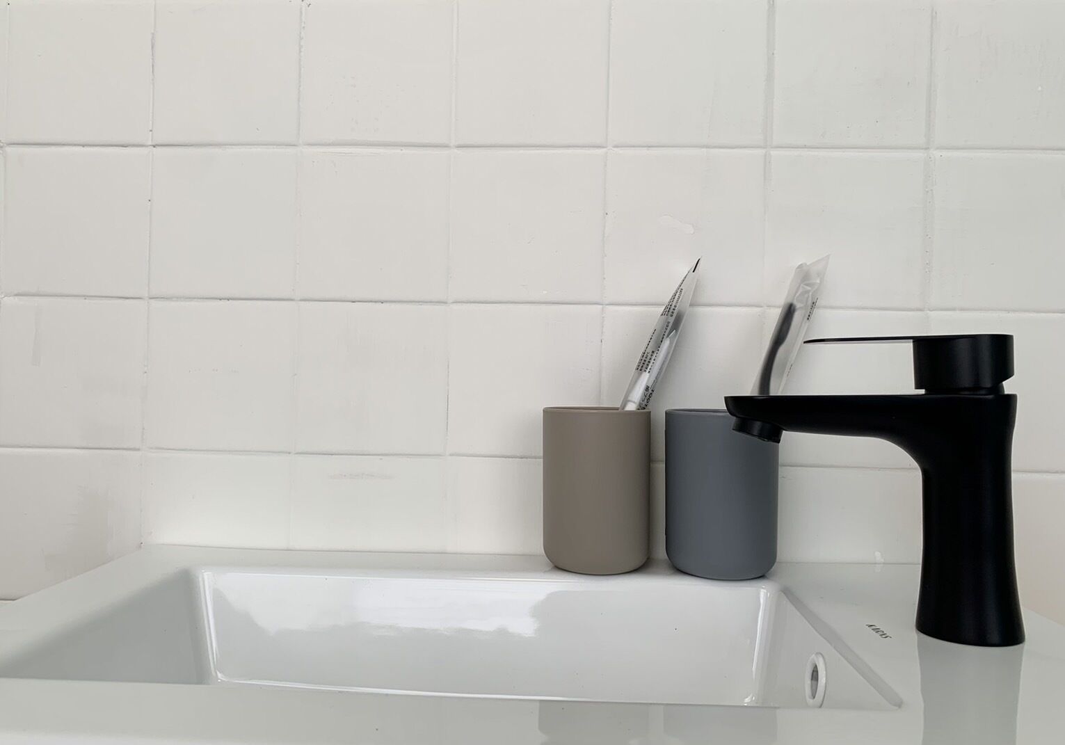 Une salle de bain avec un mur recouvert de carreaux blancs, un robinet noir et deux récipients cylindriques, l’un beige et l’autre gris, contenant des brosses à dents.