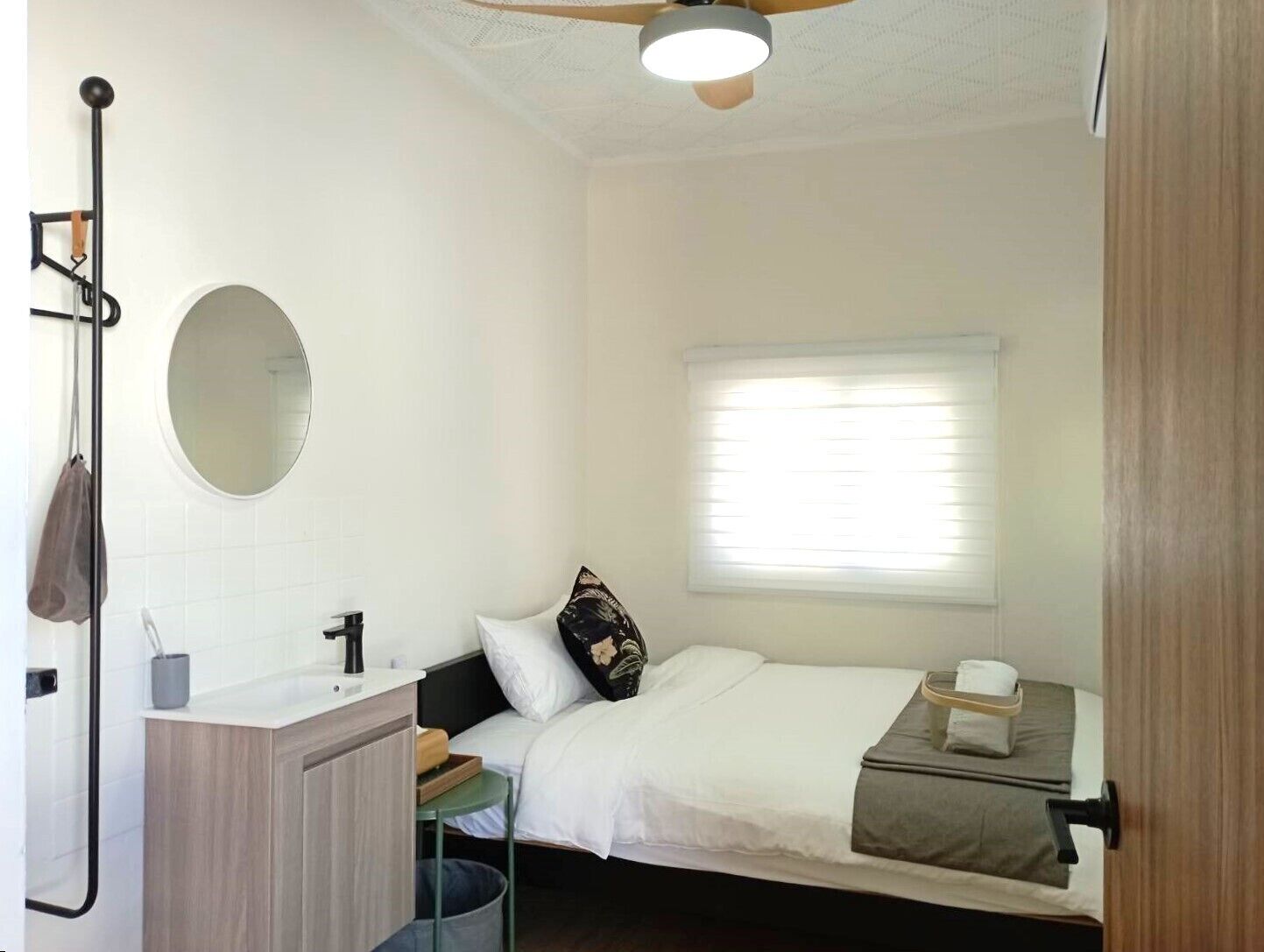 Un dormitorio compacto con una cama individual, una mesita de noche pequeña, un espejo y una ventana con persianas.