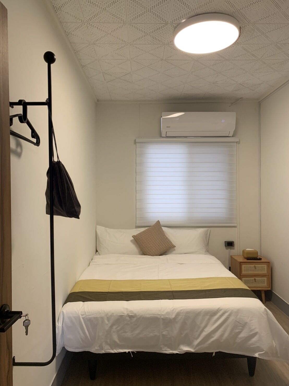 Une chambre d’hôtel équipée d’un lit, d’un porte-bagages suspendu, d’un climatiseur, d’une table de chevet et d’une fenêtre avec des stores.