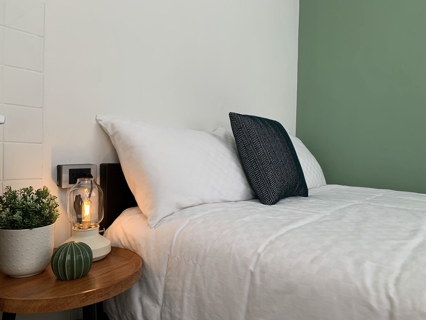 A neatly made bed with white bedding and two pillows, a small bedside table with a potted plant and a decorative item, and a wall socket.