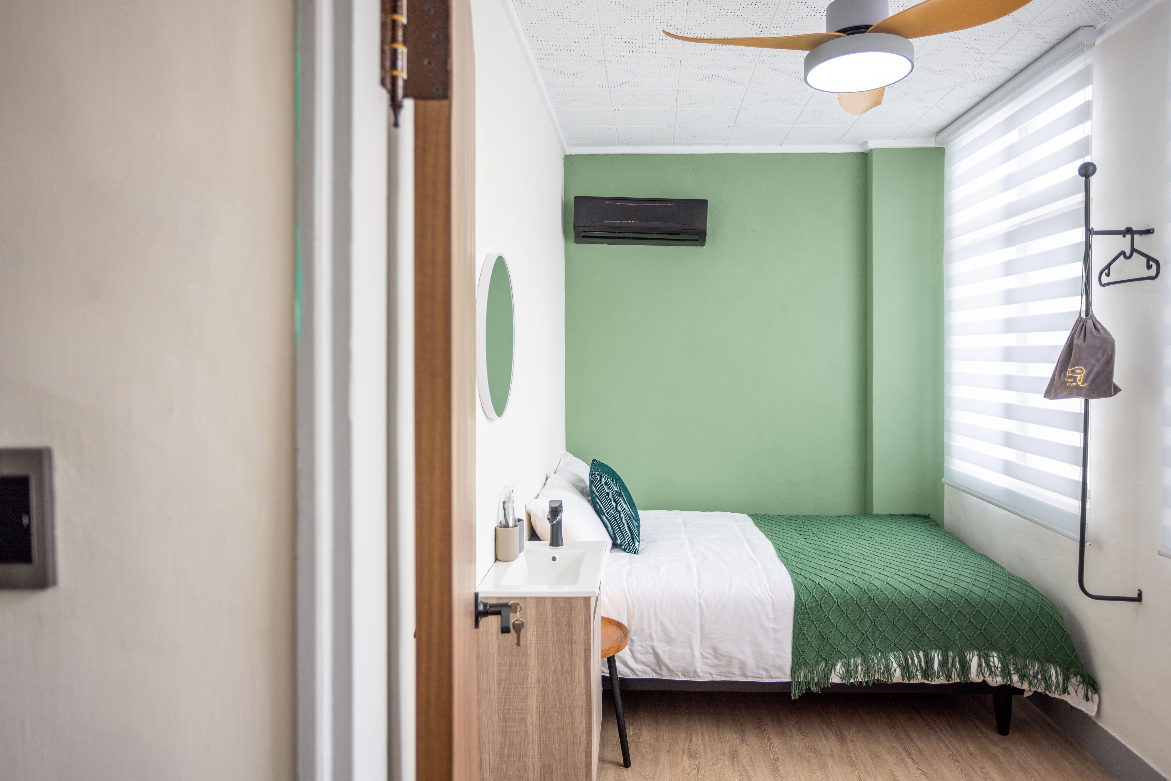 A bedroom with a bed, a bedside table, a mirror, an air conditioning unit, and a window with blinds.