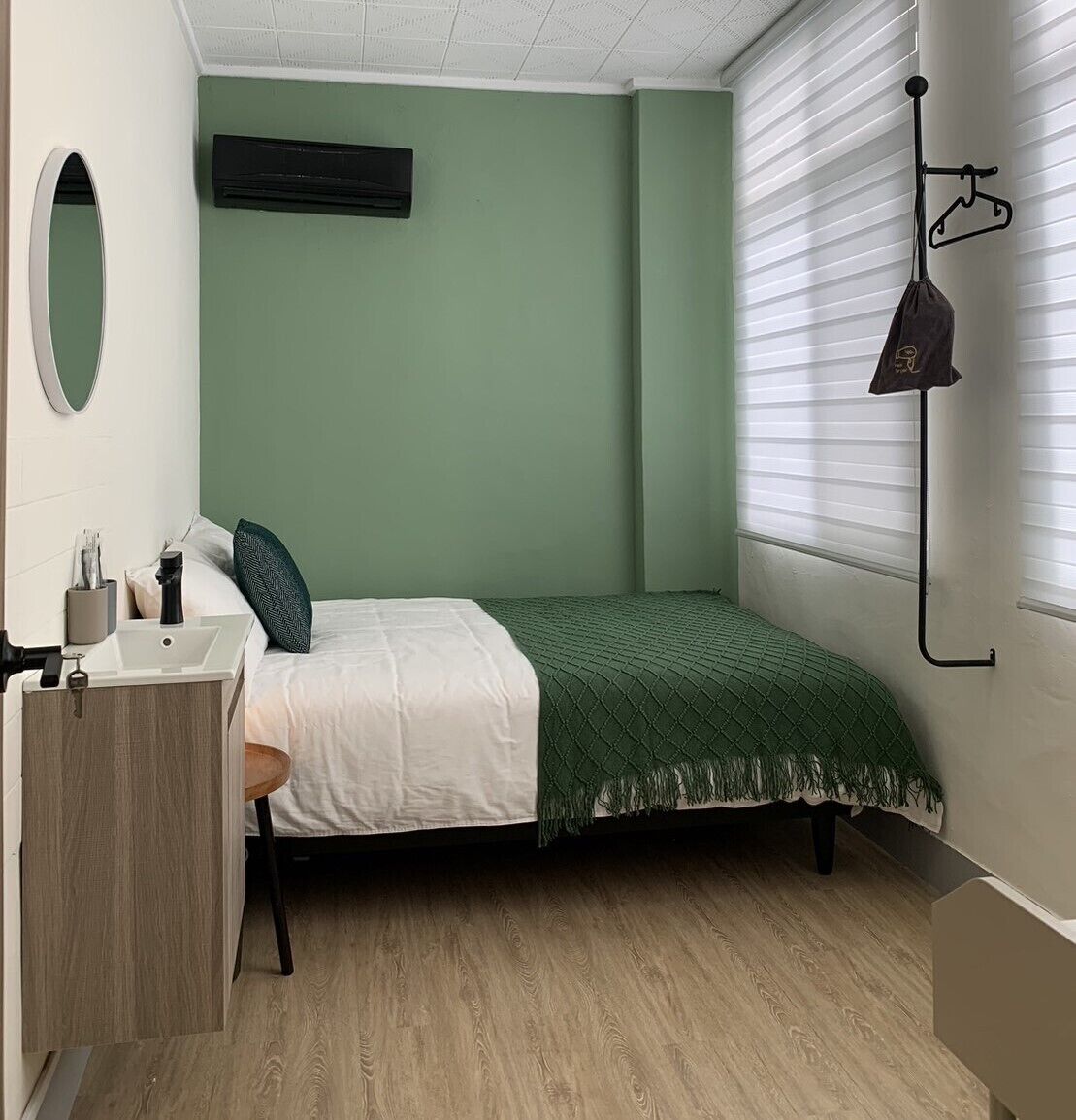A small, neatly arranged bedroom with a single bed, a bedside table with a lamp, a mirror, and a window with blinds.