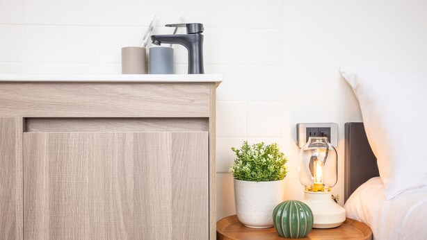 A bathroom with a wooden cabinet, a sink with a faucet, a potted plant, a green vase, and a bedside lamp.